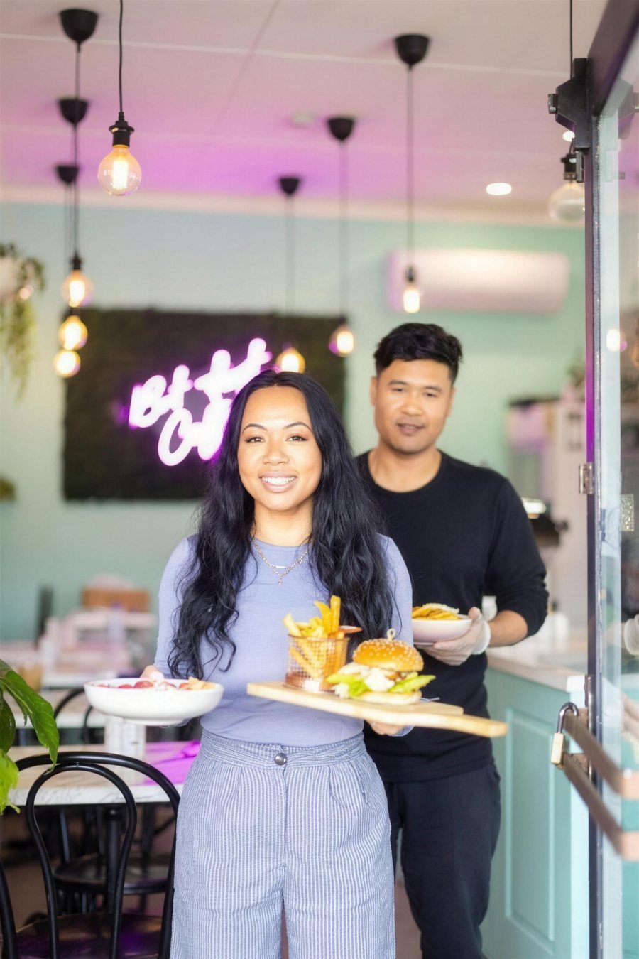 Two cafe owners holding boards of food under a neon sign